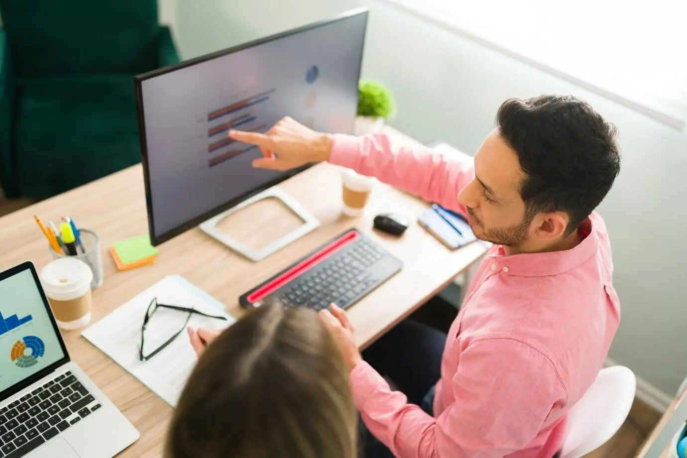 An attractive man sitting in an office points to a computer screen and a business presentation, while professional coworkers work on an analytical report.