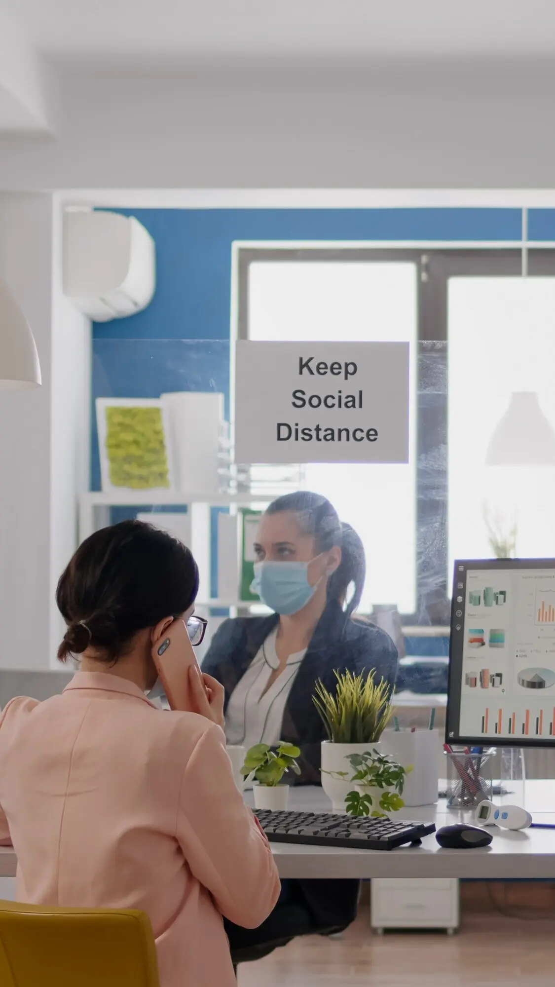 A businesswoman wearing a medical face mask is speaking on her smartphone with partners while her coworkers work...